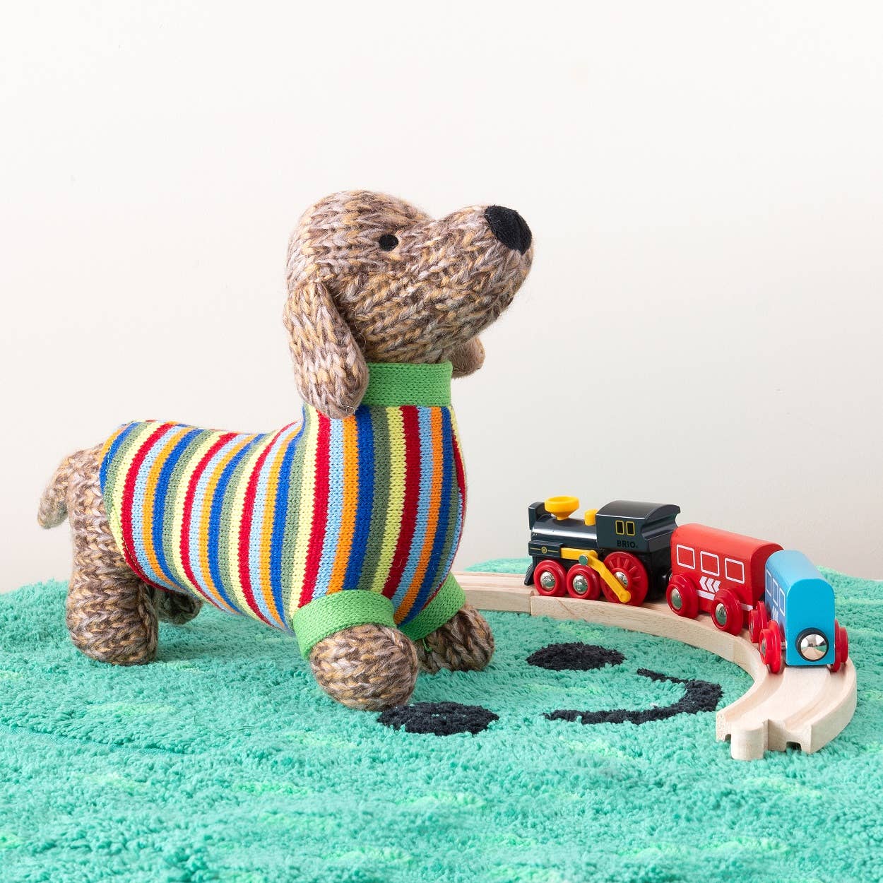 Stuffed dog in a colorful sweater with a toy train set on a green surface.