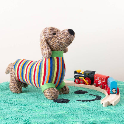 Stuffed dog in a colorful sweater with a toy train set on a green surface.
