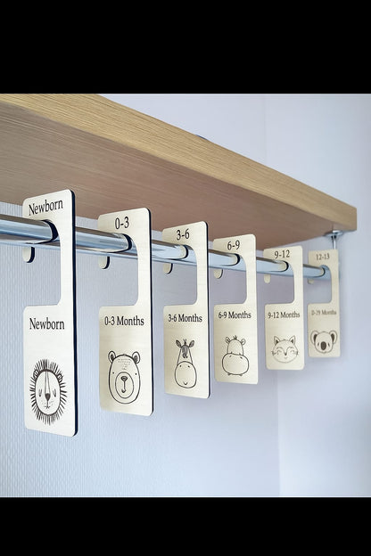 Clothing size tags labeled from Newborn to 24 Months on a wooden shelf.