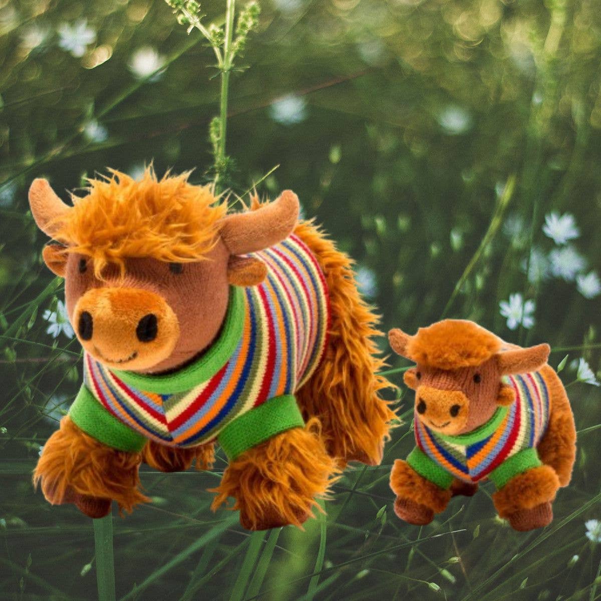 Two plush cow toys wearing colorful sweaters in a grassy outdoor setting