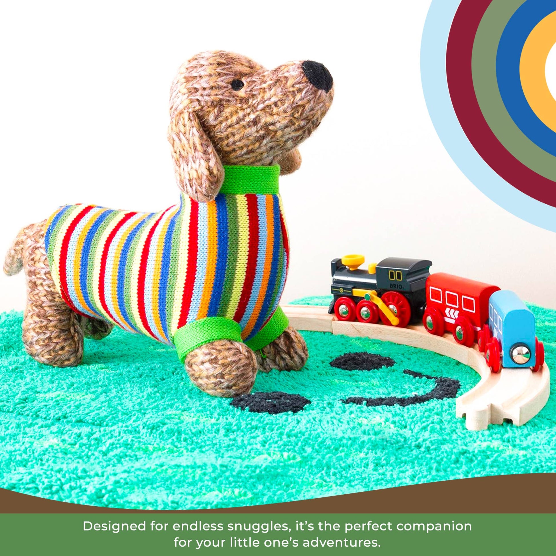 Stuffed dog toy with a colorful striped sweater near a toy train set on a green surface.