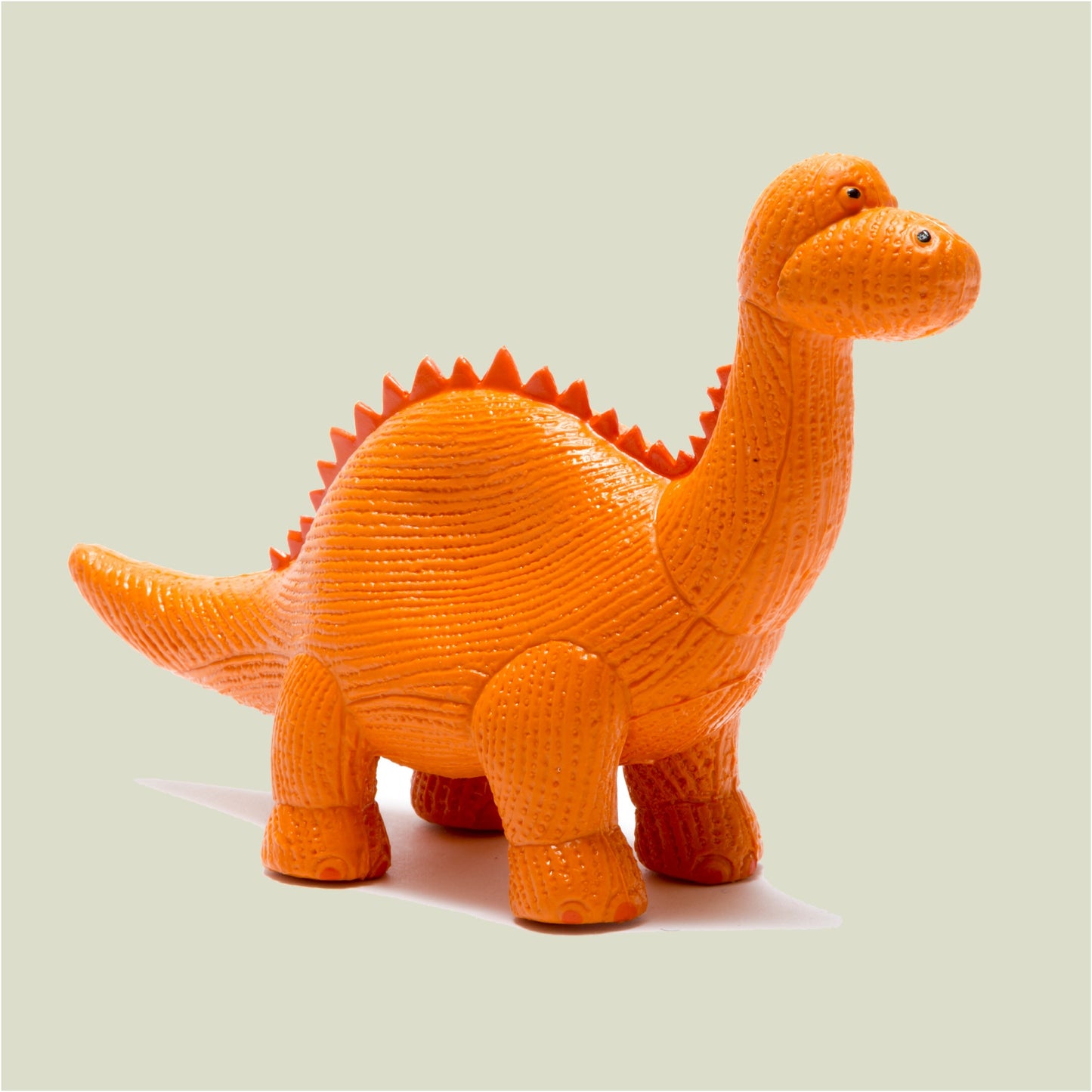 An orange textured dinosaur toy, resembling a Brachiosaurus, shown from a three-quarters angle against a solid light sage green background.